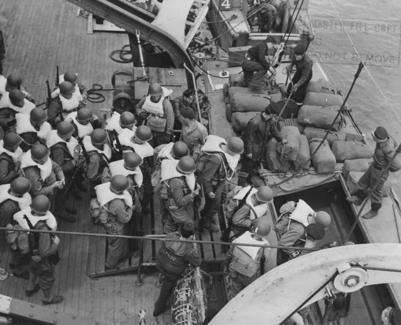 Seen from above, troops load from a ship into a small landing craft at the ship's side