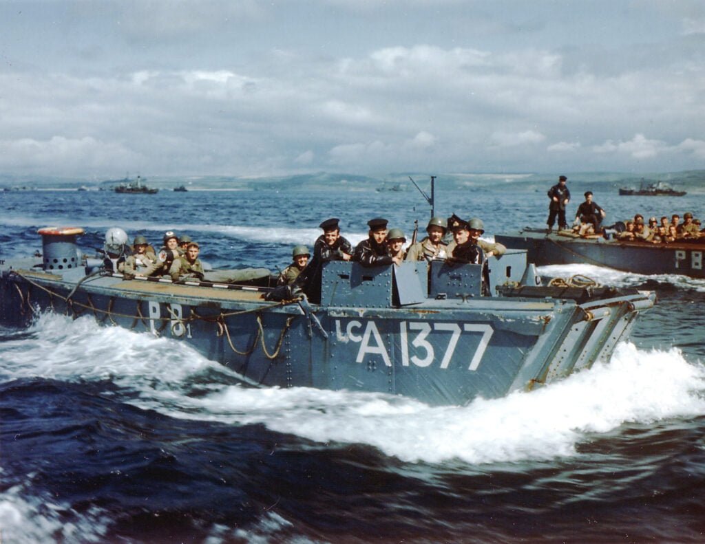 A colour photograph of a small landing craft seen from the side. It is full of troops.