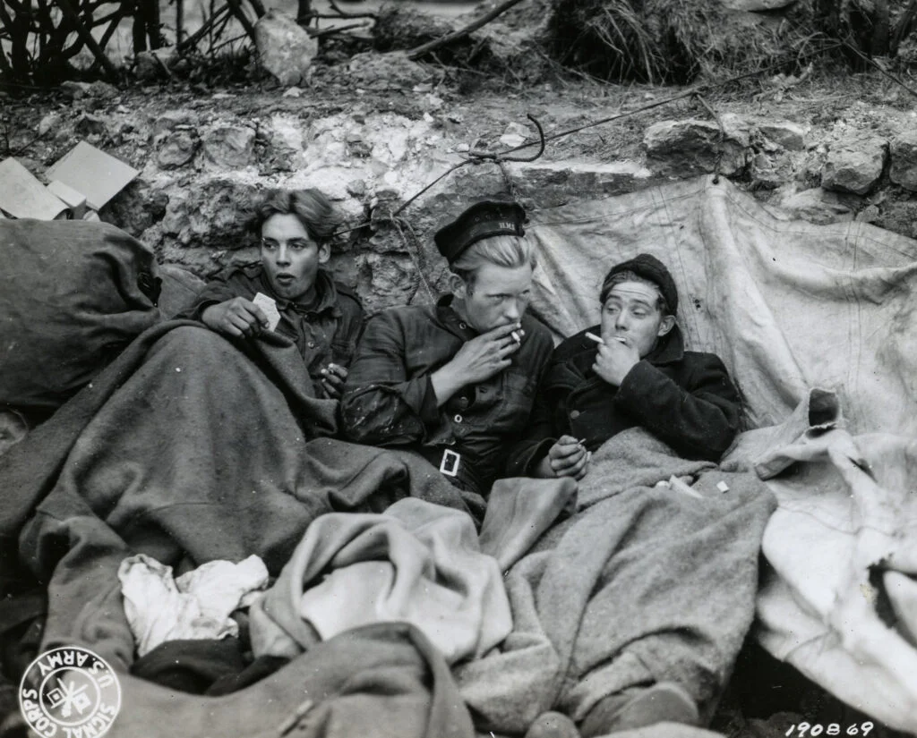 Three men lying on a beach under blankets. They look tired and are eating or smoking.