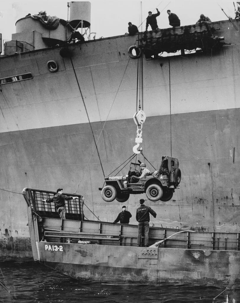 A jeep being lowered from a ship into a Landing Craft, Mechanised below