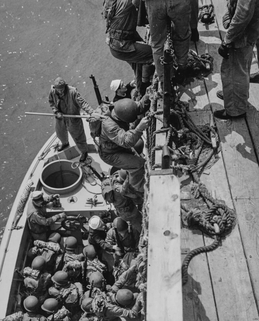 A landing craft full of men, seen from a above from the side of an adjacent ship