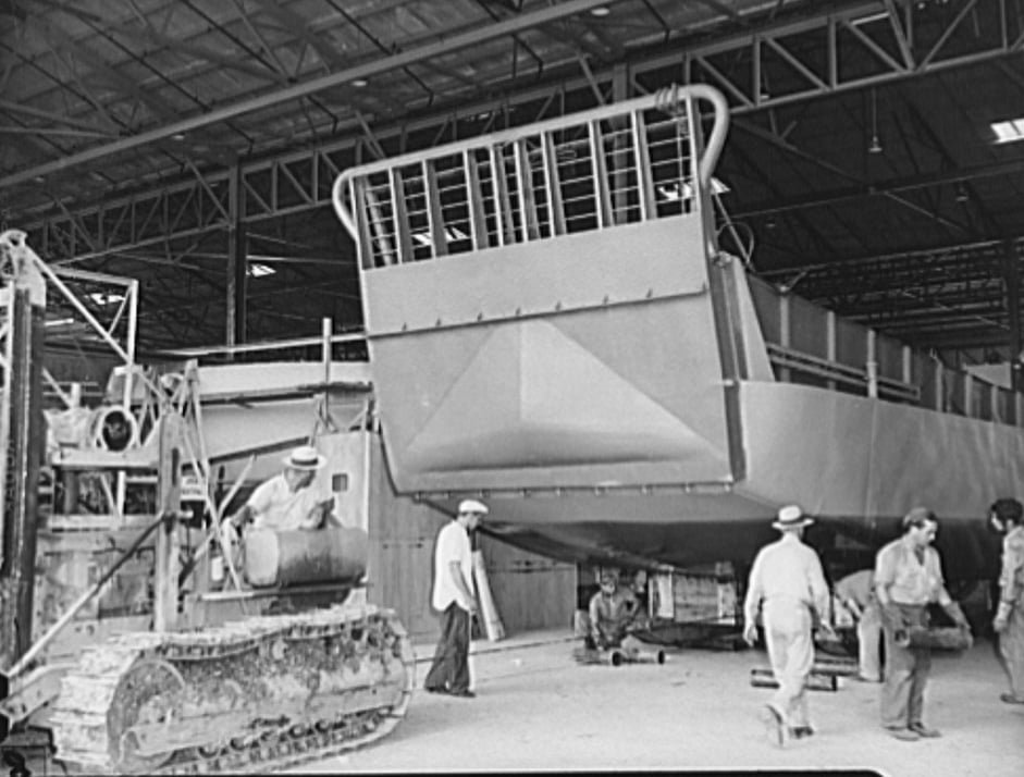 A landing craft being towed by a tracked vehicle, inside a large building
